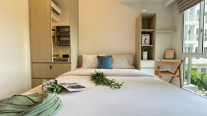 A modern hotel room with a bed, a side table, a chair, a bookshelf, and a window with a view of buildings. - Basic And Compact Studio At Tokyo Riverside Pik 2 Apartment (Teluknaga)