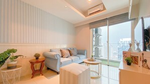 A modern living room with a sofa, a small round table, and a white ottoman. There is a large window with a view of the city. - Cozy Living 2Br Apartment Casa Grande Residence (Jakarta)