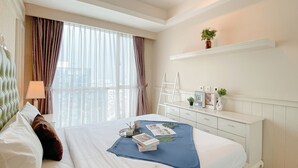 A hotel room with a large bed, a bedside table, a dresser, a window with curtains, and a view of the city. - Cozy Living 2Br Apartment Casa Grande Residence (Jakarta)