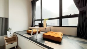 A modern room with a large window, a small table with a plant, and two orange cushions. - Studio Apartment Collins Boulevard Near Summarecon Mall Serpong (Gading Serpong)