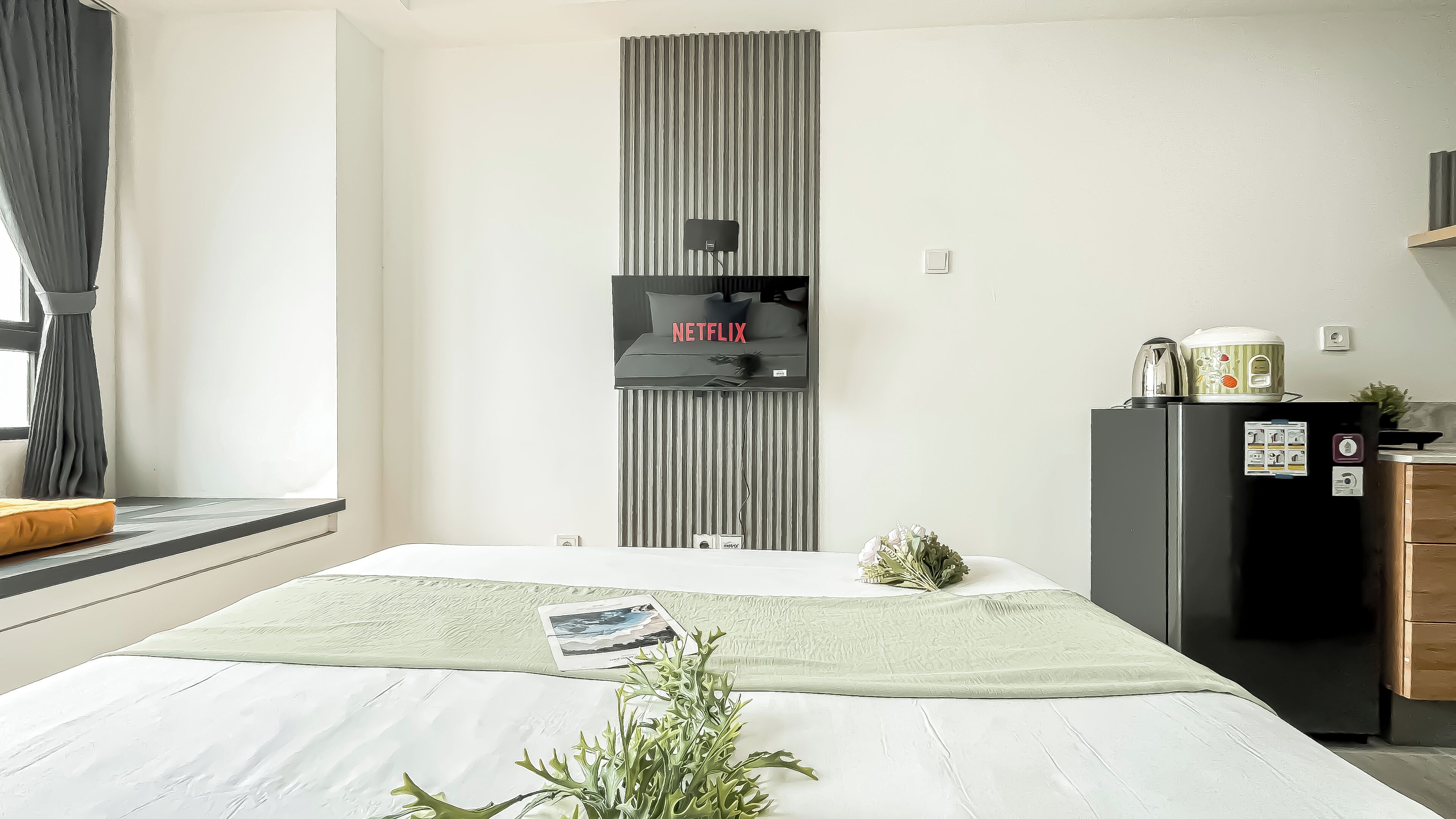 Un dormitorio moderno con una cama, un televisor de pared, un refrigerador y una ventana con cortinas.
