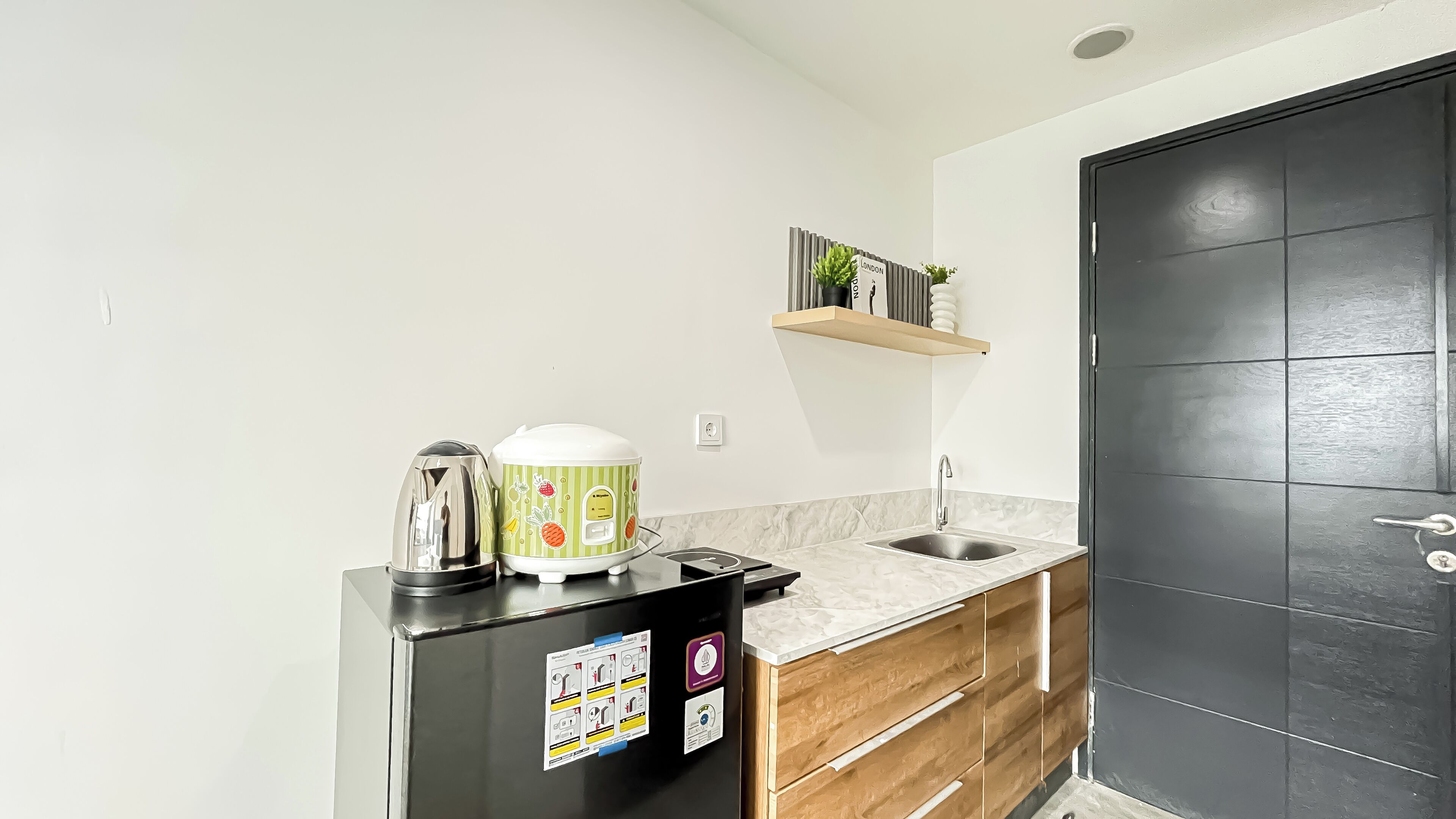 A modern kitchen with a stainless steel refrigerator, a rice cooker, and a sink. There is a wooden cabinet below the countertop and a black door to the right.