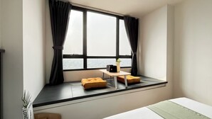 A room with a large window, a small table with a plant, and two yellow cushions. - Studio Apartment Collins Boulevard Near Summarecon Mall Serpong (Gading Serpong)
