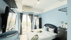 A hotel room with a bed, a small table, a chair, a window with curtains, an air conditioning unit, and a view of the outside. - Studio Apartment Poris 88 Near Batu Ceper Station (Tangerang)