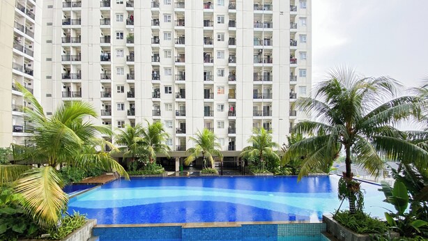 Une piscine dont le bord est revêtu de carreaux, entourée de palmiers et de verdure, avec un immeuble de grande hauteur en arrière-plan.