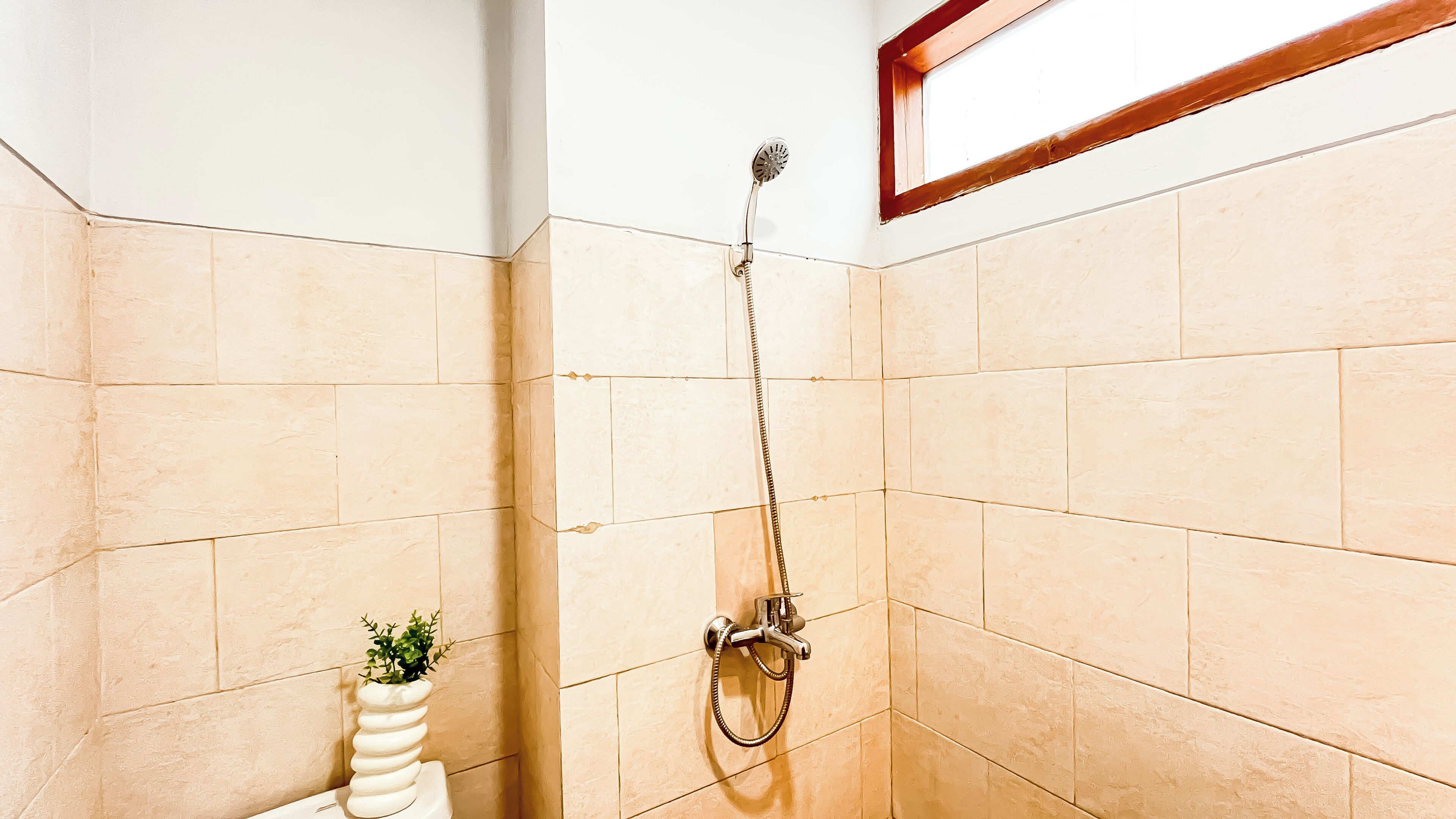 A bathroom with beige tiles, a shower with a handheld showerhead, and a small potted plant.