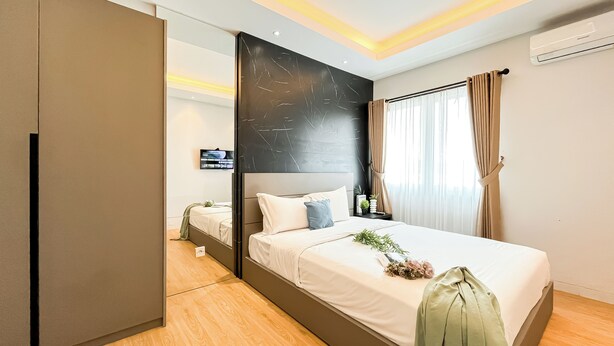 Una habitación de hotel moderna con una cama grande, un televisor de pantalla plana, una mesita de noche con una planta y una ventana con cortinas.