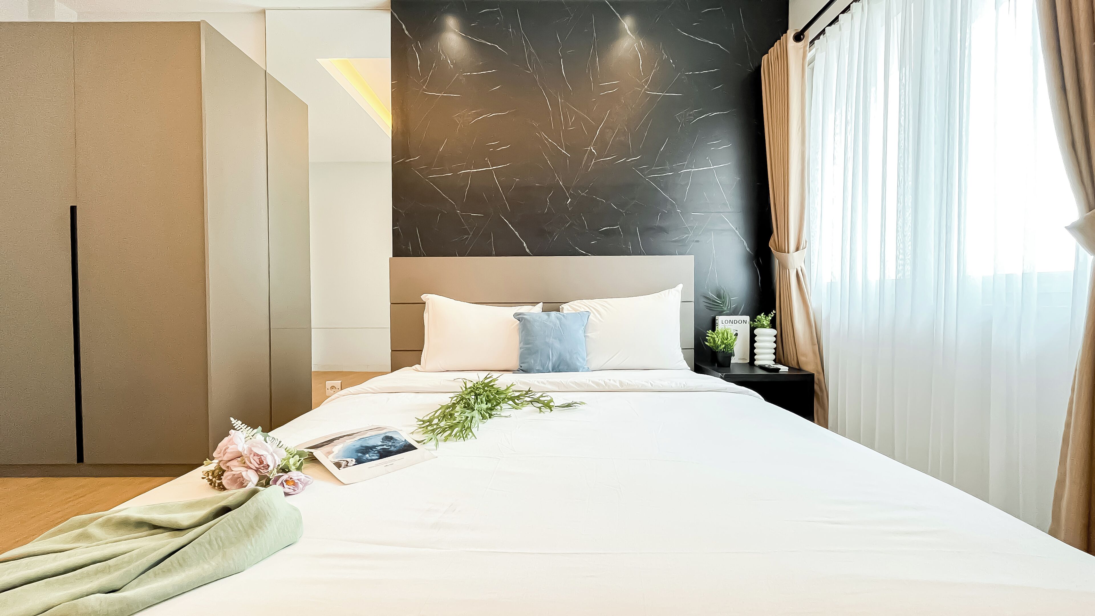 A modern hotel room with a large bed, a nightstand, a potted plant, and a view of the city.