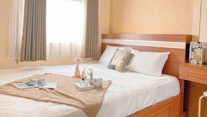 A neatly made bed with a beige blanket, a bedside table with a book and a bottle, and a window with curtains. - Simple Style 2Br At Green Pramuka City Apartment (Jakarta)