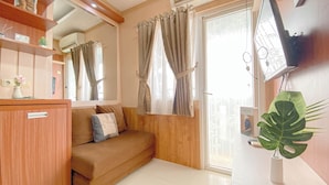 A compact living space with a sofa, a small cabinet, a plant, and a television mounted on the wall. - Simple Style 2Br At Green Pramuka City Apartment (Jakarta)
