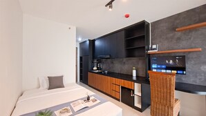 Une chambre d’hôtel moderne équipée d’un lit, d’une petite kitchenette, d’un téléviseur fixé au mur et d’un fauteuil en osier.