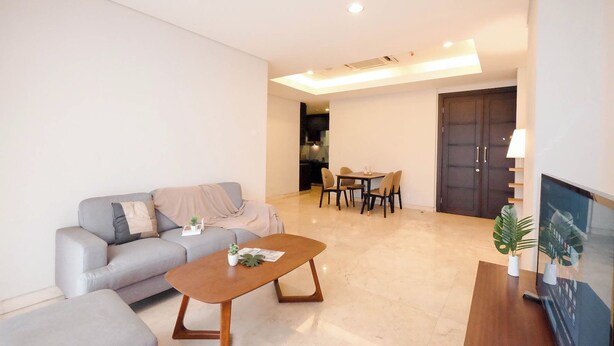 A modern living room with a grey sofa, a wooden coffee table, and a dining area with a table and chairs. - Strategic Place 2Br Apartment With Private Lift Masterpiece The Epicentrum Kuningan (Jakarta)