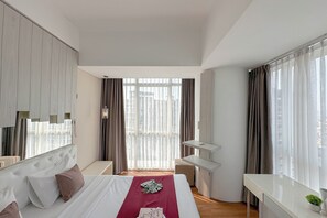 A modern hotel room with a large bed, a desk, and a view of the city through sheer curtains. - Modern Minimalist 2Br At Linden Apartment (Surabaya)