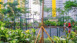 An outdoor gym area with exercise equipment, surrounded by lush greenery and tall buildings in the background.