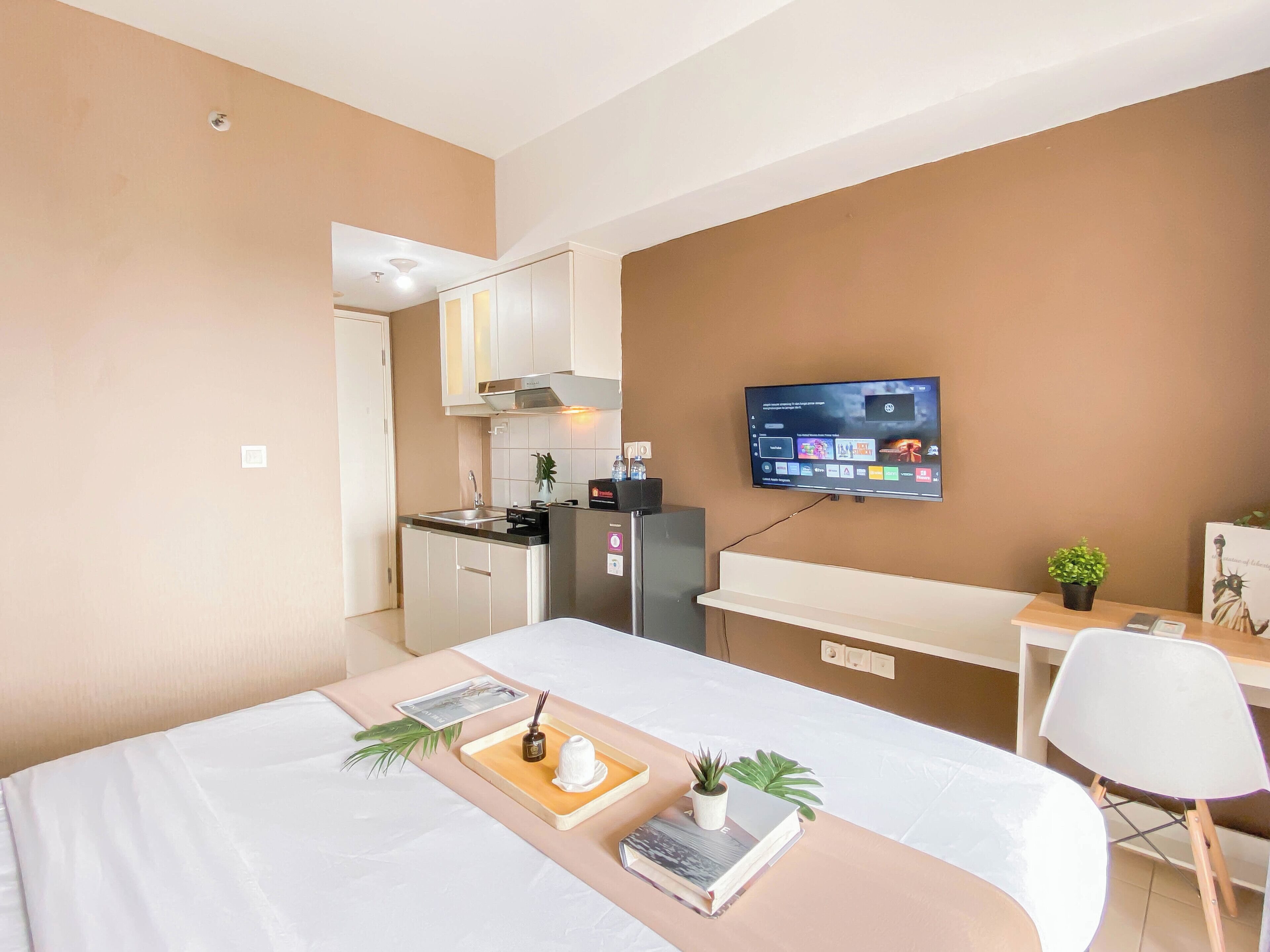 A modern hotel room with a bed, bedside table, a small desk with a chair, a TV mounted on the wall, and a kitchenette area.
