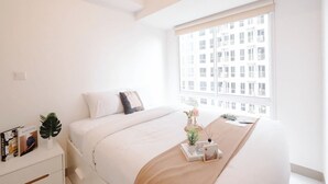 Un dormitorio minimalista con un ventanal grande, cama con ropa de cama blanca, mesita de noche con libros y una planta, y vista a un edificio exterior.