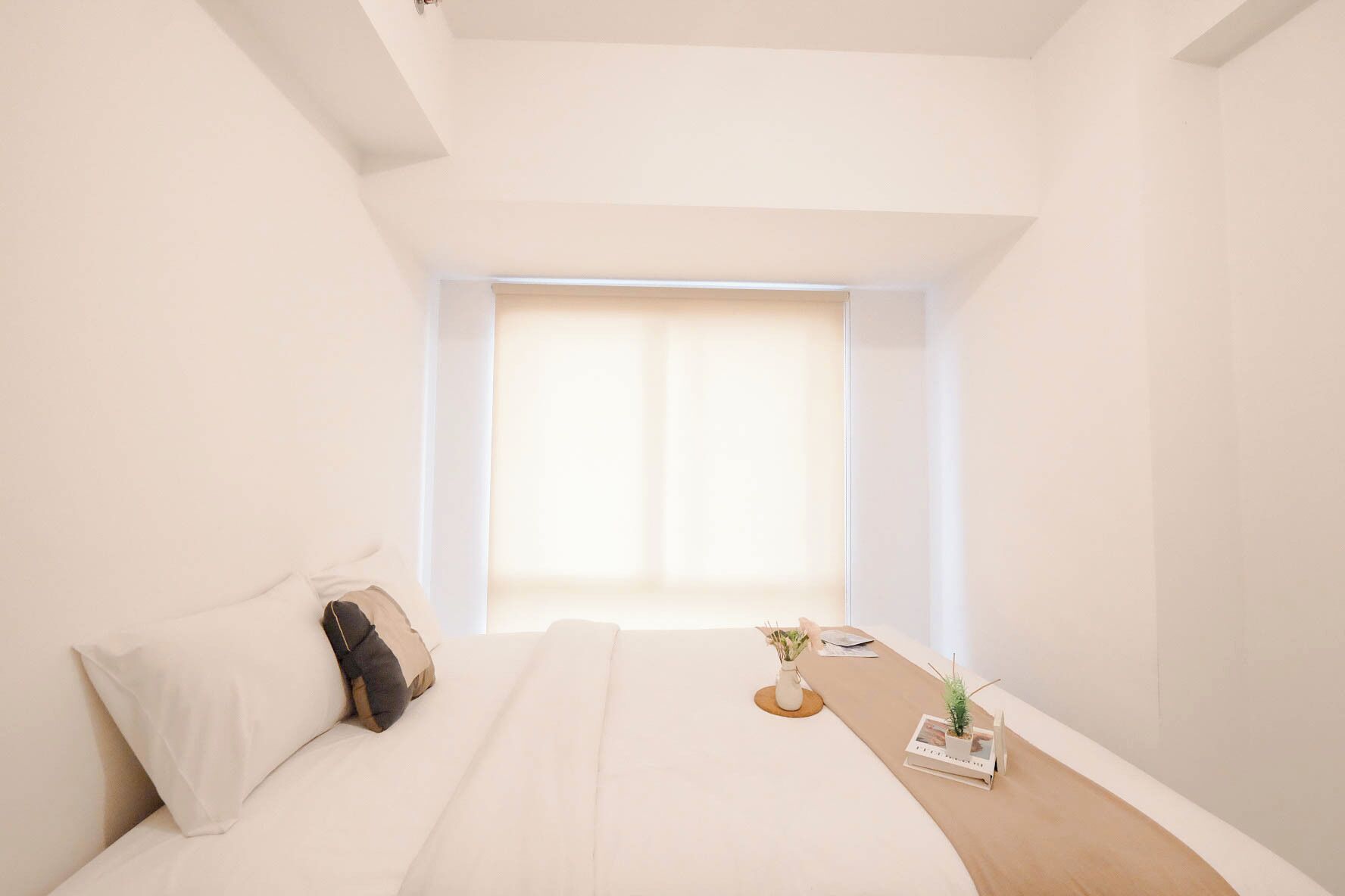 A minimalist hotel room with a bed, a small table with a plant, and a window with curtains.