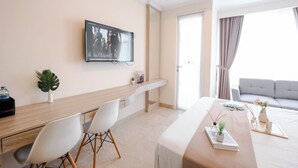 A modern hotel room with a wooden desk, white chairs, a flat-screen TV, a sofa, and a large window with curtains. - Modern Studio Near Taman Ismail Marzuki At Apartment Menteng Park (Jakarta)