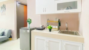 A modern kitchen with white cabinets, a built-in dishwasher, a sink, and a small countertop with a plant and a dish rack. - Soft Pastel Minimalist 3Br At Pollux Chadstone Apartment (Cikarang)