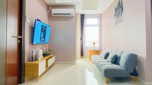 A living room with a flat-screen TV, a sofa, a small table with a vase, and an air conditioning unit on the wall. - Soft Pastel Minimalist 3Br At Pollux Chadstone Apartment (Cikarang)