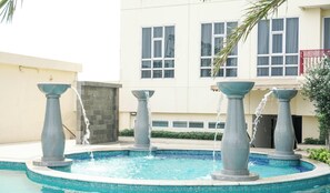 A swimming pool with four fountains, surrounded by a tiled deck and a building with large windows and a balcony. - Boho Touch 2Br Apartment With Extra Room At Lucky Tower Residence (Jakarta)
