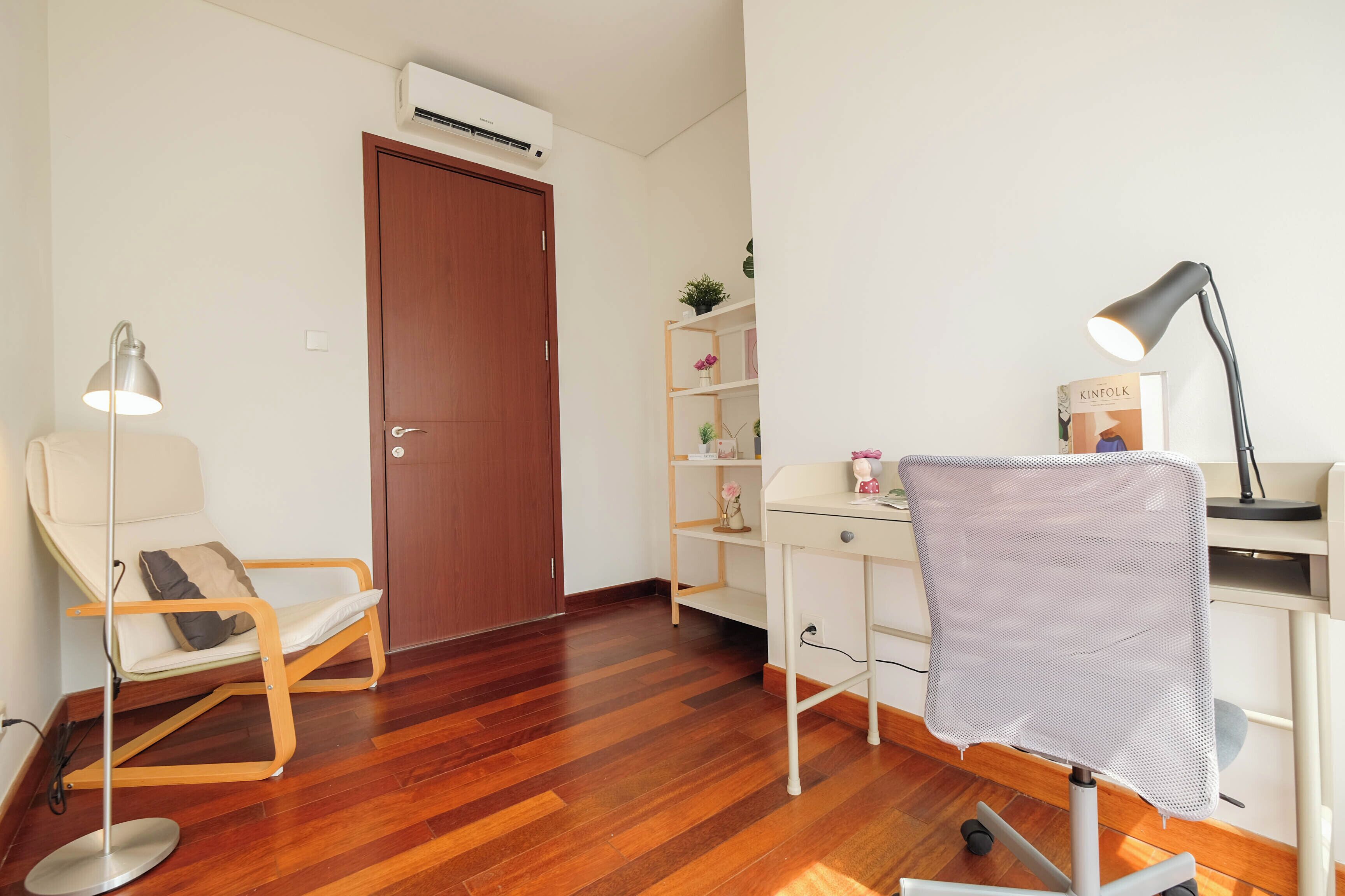 A room with a wooden floor, a desk with a chair, a lamp, and a bookshelf.