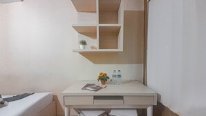 Une petite pièce, bien rangée, avec un bureau, un lit et une étagère fixée au mur, sur laquelle se trouvent une plante et deux bouteilles.
