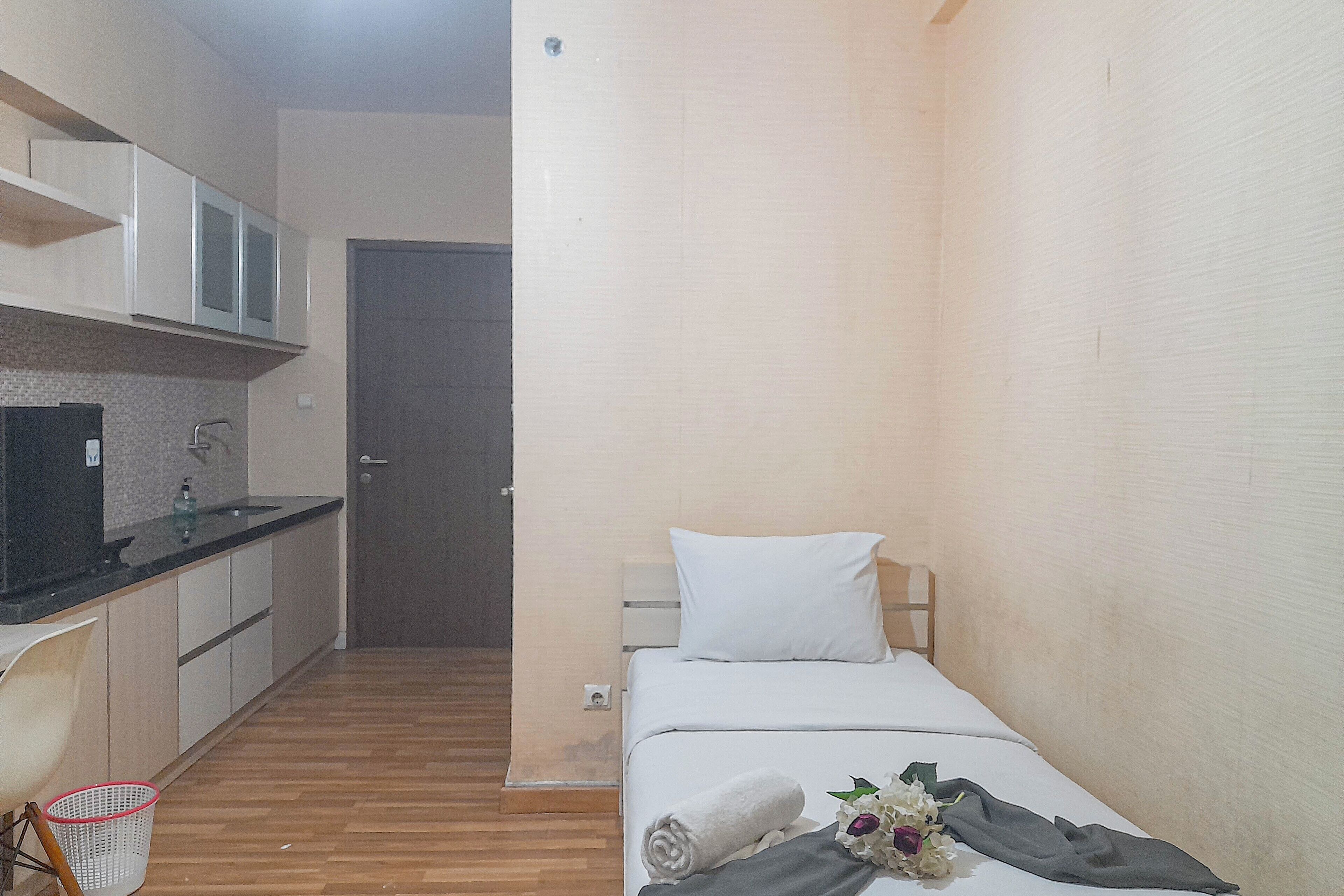 Habitación de hotel compacta con kitchenette, una cama con almohada blanca y una mesita de noche con una toalla y flores.