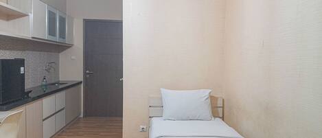 Habitación de hotel compacta con kitchenette, una cama con almohada blanca y una mesita de noche con una toalla y flores.