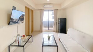 A hotel room with a flat-screen TV mounted on the wall, a sofa, a coffee table, a refrigerator, and a sliding glass door leading to a balcony.