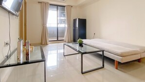 A modern room with a glass coffee table, a sofa, a flat-screen TV mounted on the wall, a black refrigerator, and a sliding glass door leading to a balcony.