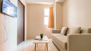A modern living room with a flat-screen TV mounted on the wall, a sofa, a coffee table with a plant, and a window with curtains. - Simple 2Br With Extra Room Transpark Juanda Bekasi Timur Apartment (Bekasi)