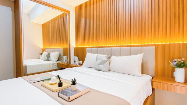 A modern hotel room with a large bed, a bedside table with a lamp, a mirror, and a wooden headboard. - Simple 2Br With Extra Room Transpark Juanda Bekasi Timur Apartment (Bekasi)