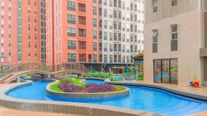 A swimming pool with a bridge, surrounded by greenery and a multi-story building with balconies. - Simple 2Br With Extra Room Transpark Juanda Bekasi Timur Apartment (Bekasi)