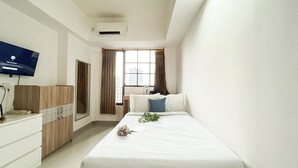 A hotel room with a bed, a TV mounted on the wall, a window with curtains, and a small table with a plant.