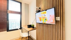A modern office space with a large flat-screen TV mounted on a wooden panel wall, a desk with a chair, and a window with blinds. - Wooden Nest 2Br Without Living Room At Evenciio Margonda Apartment (Depok)