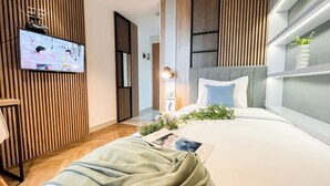 A modern hotel room with a wooden accent wall, a flat-screen TV, a bed with a blue pillow, and a bedside table with a plant. - Wooden Nest 2Br Without Living Room At Evenciio Margonda Apartment (Depok)