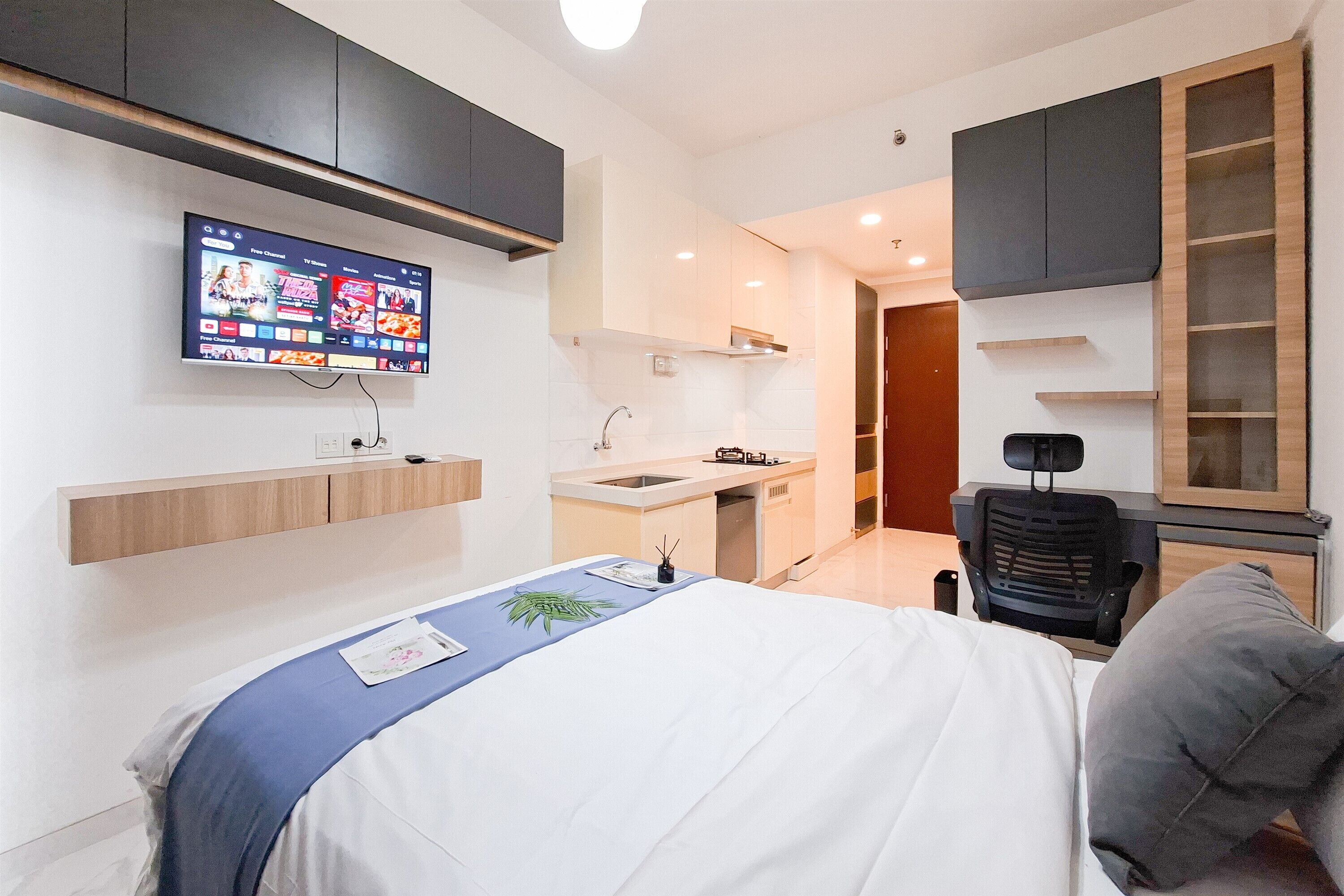 A modern hotel room with a bed, a desk, a chair, a TV mounted on the wall, and a kitchenette.