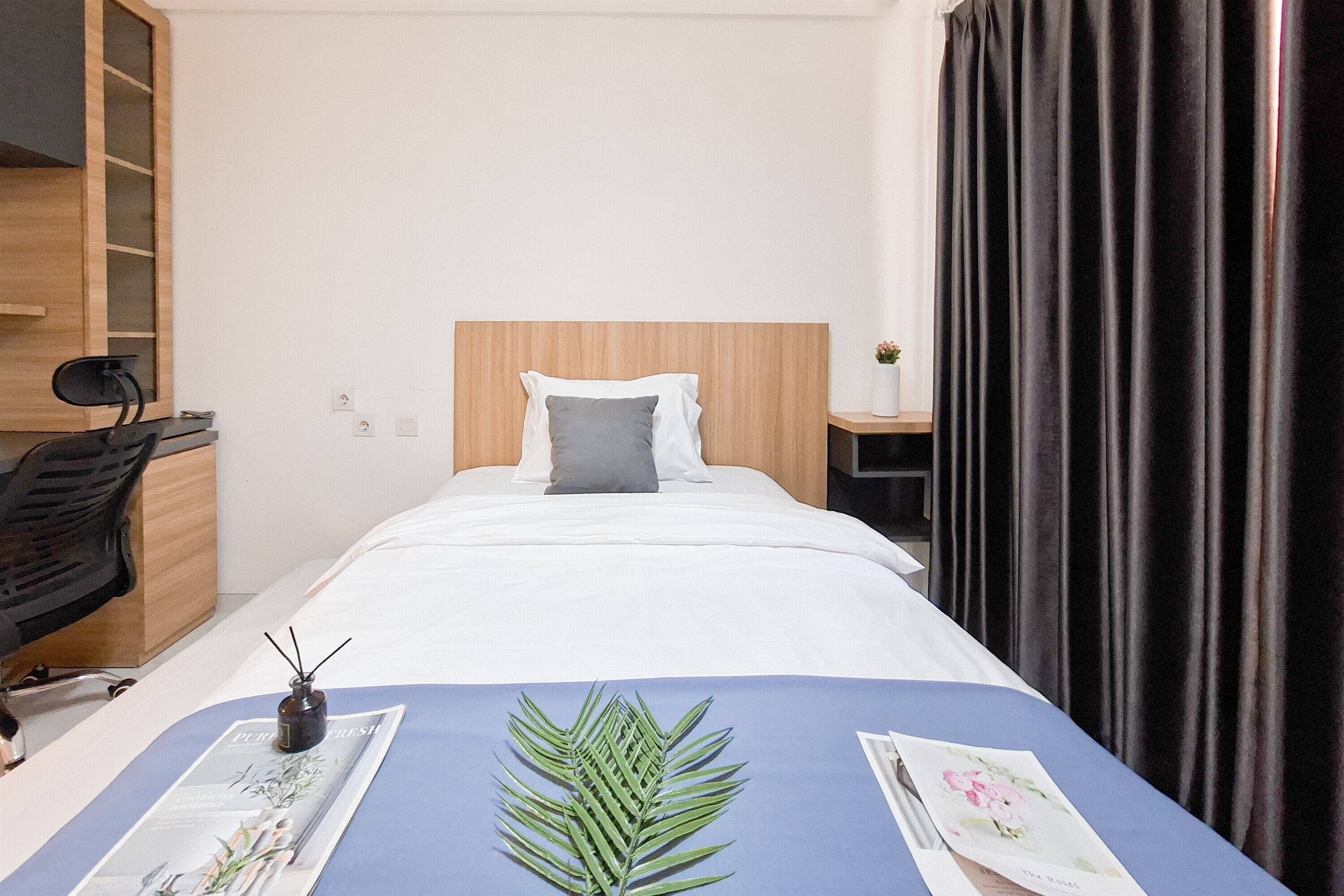 A hotel room with a wooden headboard, a bed with white linens, a blue bedspread, a black chair, a small table with a vase, and a dark curtain.