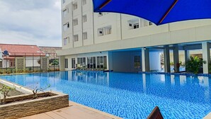 En swimmingpool med en kakelbeklädd kant, omgiven av en byggnad med stora fönster och ett blått solskydd.