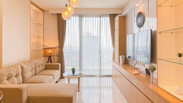 A modern living room with a sofa, a small table, a lamp, a TV, and a view of a cityscape through sheer curtains. - Warm Contemporary 2Br Apartment At Samara Suites (Jakarta)