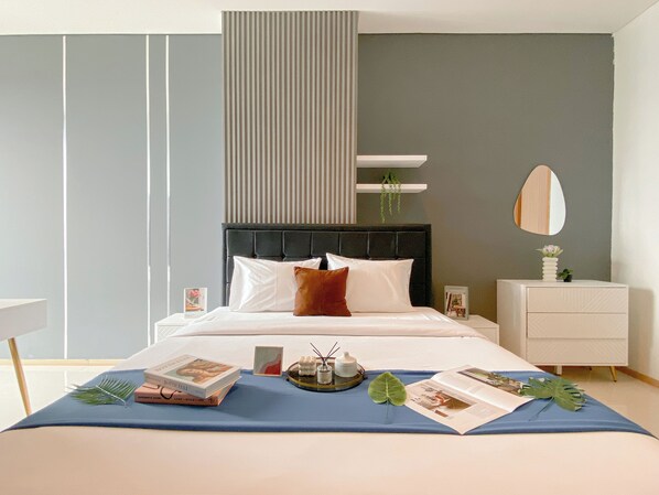 A modern bedroom with a large bed, a bedside table with a book and a vase, a dresser, and a mirror on the wall. - Modern Monochromatic 2Br Near Lrt Station At Apartment Samara Suites (Jakarta)