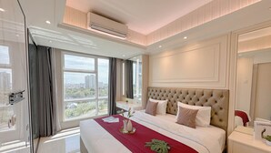 A hotel room with a large bed, a bedside table, a nightstand, a window with curtains, and an air conditioning unit. - Cozy And Spacious 1Br At Grand Sungkono Lagoon Apartment (Surabaya)