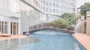 A modern hotel with a swimming pool, a stone bridge, and a view of a high-rise building. - Cozy And Spacious 1Br At Grand Sungkono Lagoon Apartment (Surabaya)