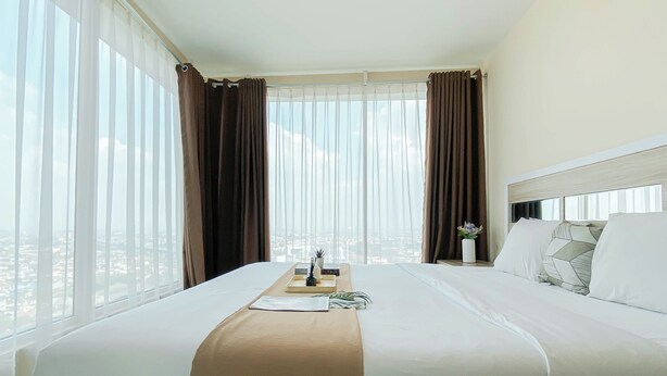 A hotel room with a large bed, a bedside table with a vase, and a view of the city through sheer curtains. - Simple And Homey 2Br At Grand Kamala Lagoon Apartment (Bekasi)