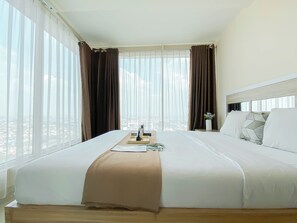 Ein Hotelzimmer mit einem großen Bett, einem Nachttisch mit einer Vase und Blick auf die Stadt durch leichte Vorhänge.