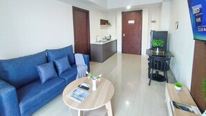 A modern hotel room with a blue sofa, a small round table with a book and a vase of flowers, a TV on a stand, and a kitchen area with a sink and countertop.