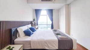 A hotel room with a bed, a bedside table with a plant, a window with curtains, and a view of the outdoors.
