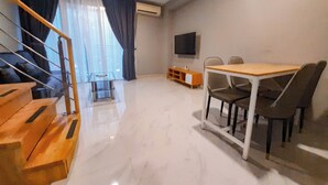 A modern living room with a wooden staircase, a dining area with a table and chairs, a TV mounted on the wall, and a large window with curtains. - Cozy Loft 2Br At Grand Jati Junction Apartment (Medan)