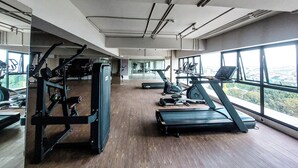 A gym with exercise equipment, large windows, and a view of the city. - Cozy Loft 2Br At Grand Jati Junction Apartment (Medan)
